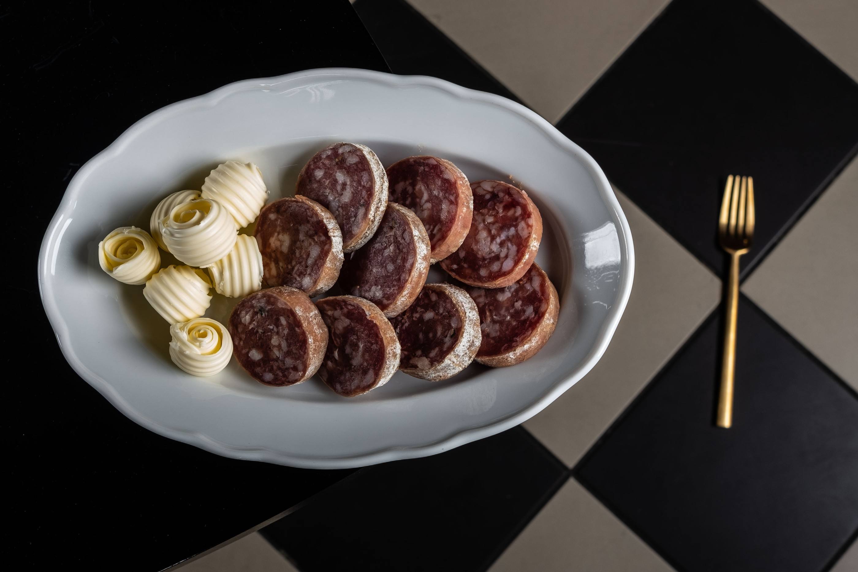 Fette di Salame della Rosa con riccioli di burro su un piatto ovale, forchetta dorata, tavolo a scacchi